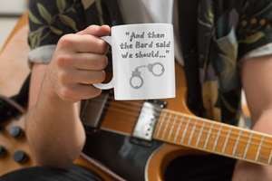 A man sitting down with an electric guitar under his arm is holding a mug that says "And then the Bard said we should..." with the image of a pair of handcuffs.