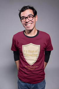 A smiling man with glasses, his hands tucked into his back jeans pockets. He is wearing a long sleeved black shirt underneath a maroon t-shirt with a tan shield that reads "+5 Shirt of Groveling" under the shield it says "(Please don't kill my character)"